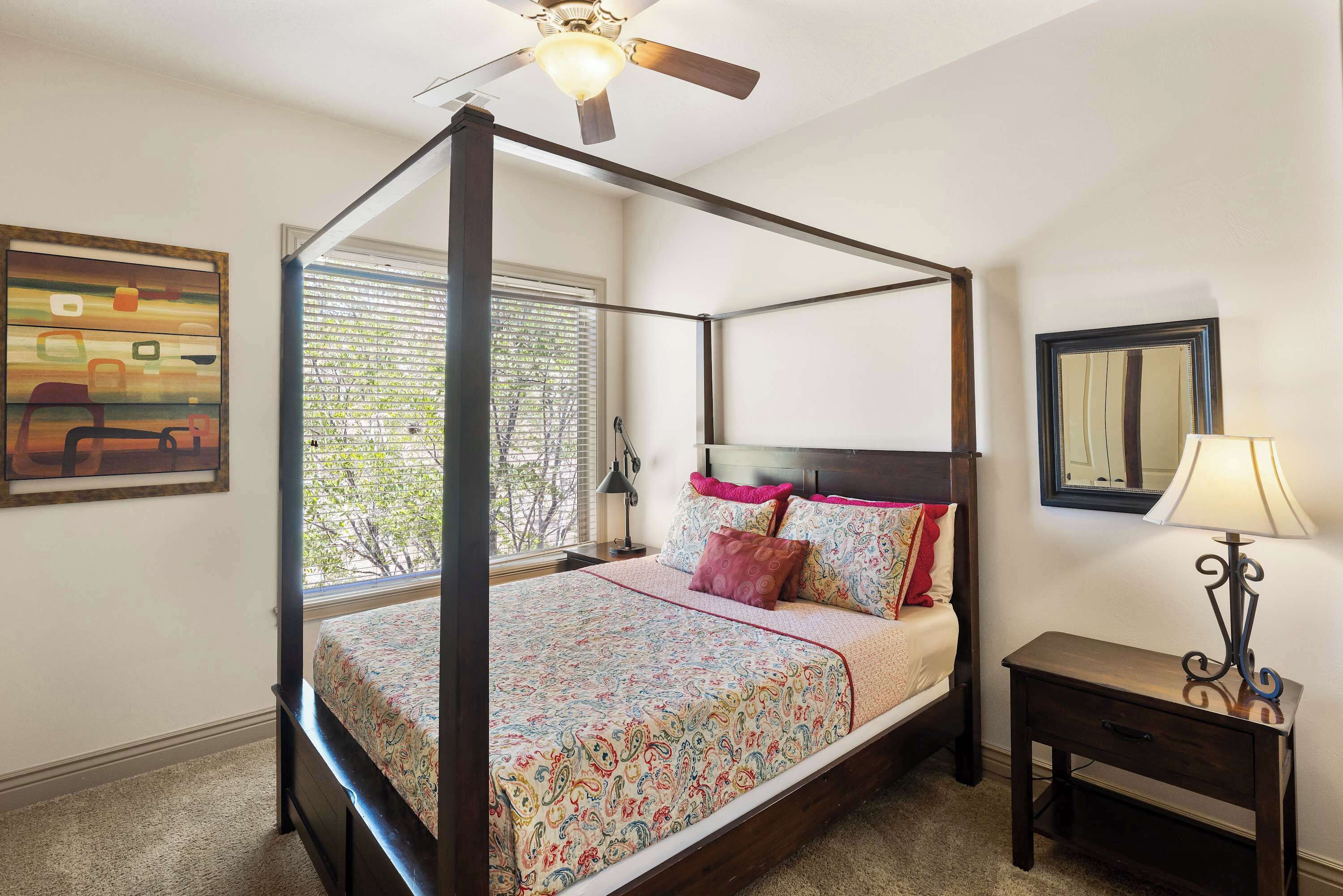 A four-poster bed with a floral bedspread and multiple pillows, a bedside table with a lamp, a ceiling fan, and a window with blinds.