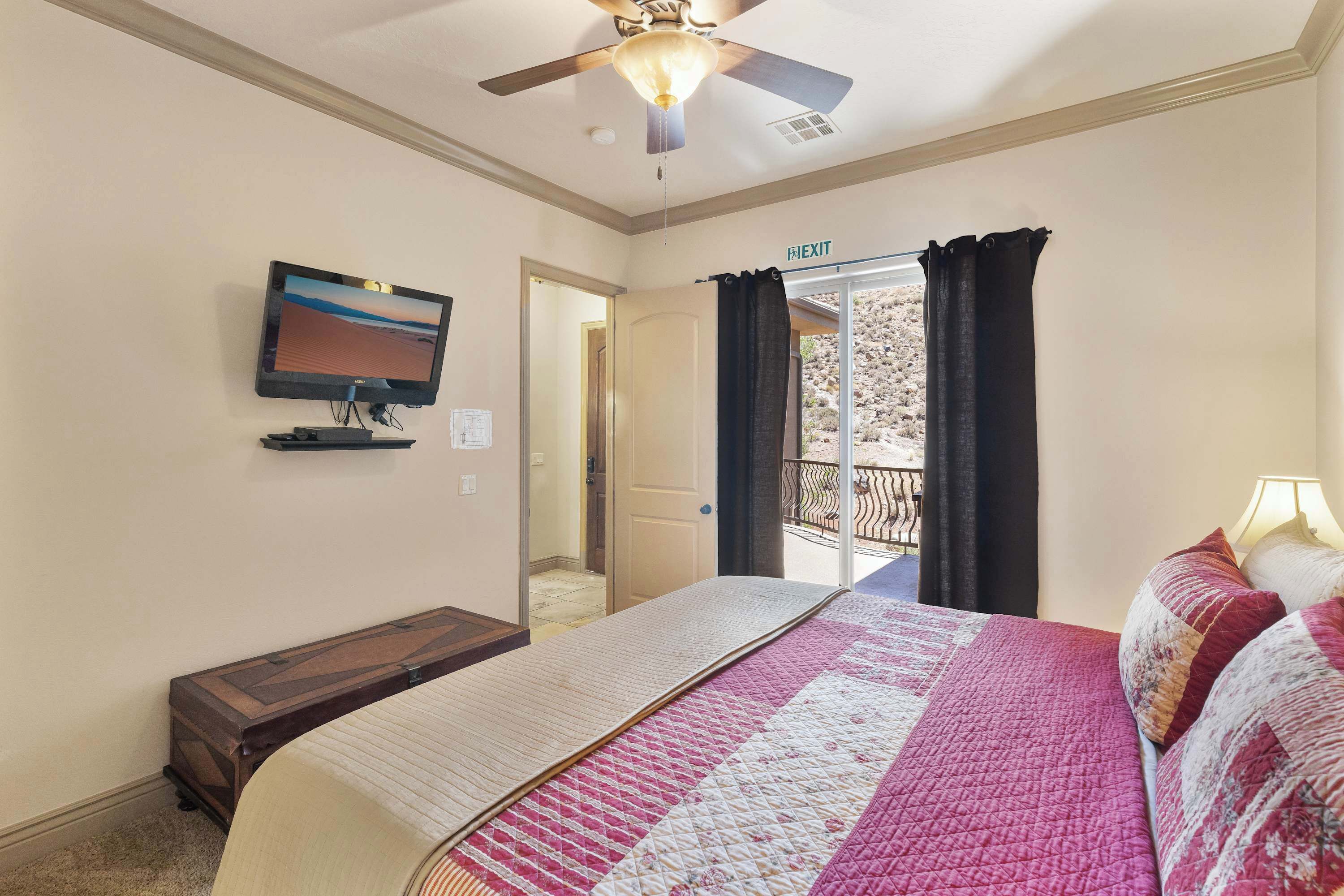 Une chambre d’hôtel équipée d’un lit, d’une télévision, d’un ventilateur de plafond et d’un balcon avec des rideaux.