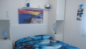 A bedroom with a bed featuring a large blue floral bedspread, white walls, and a painting of a coastal scene above the bed. - Adorable Seaview Flat at Grado Pineta (Grado)