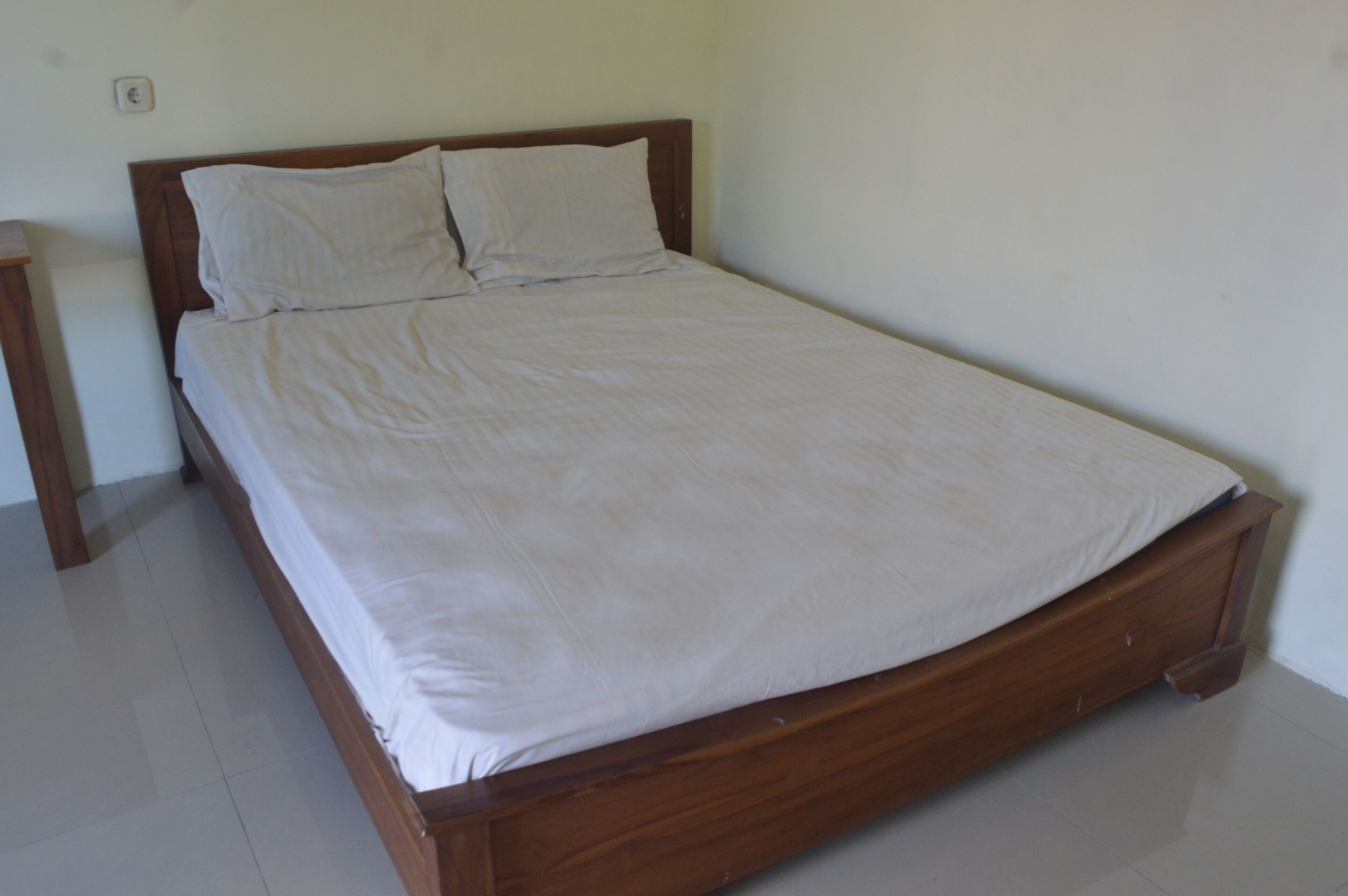 A single bed with a wooden frame and white bedding in a room with white walls and a tiled floor.