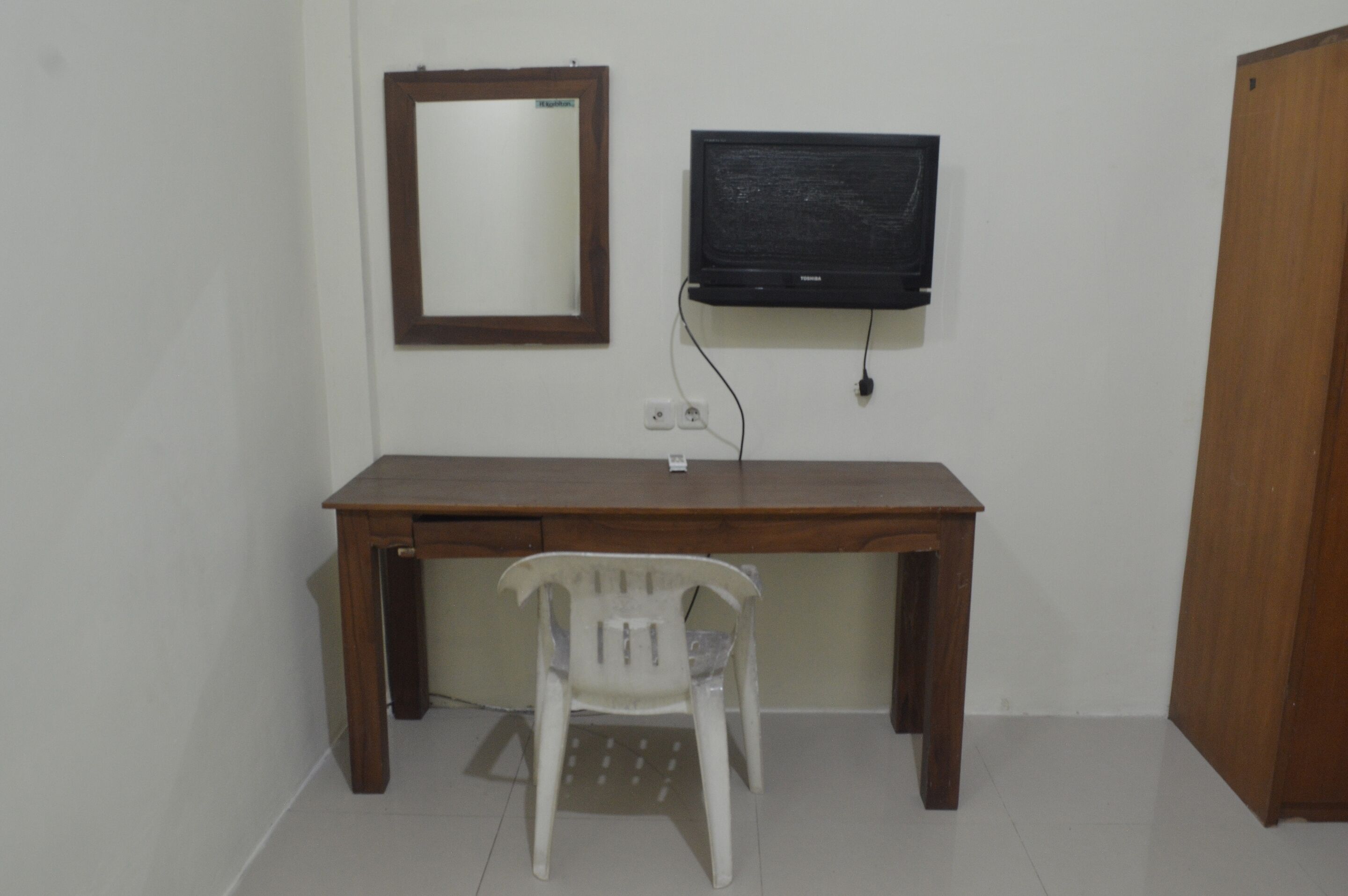 A room with a wooden desk, a plastic chair, a television mounted on the wall, and a wooden door.