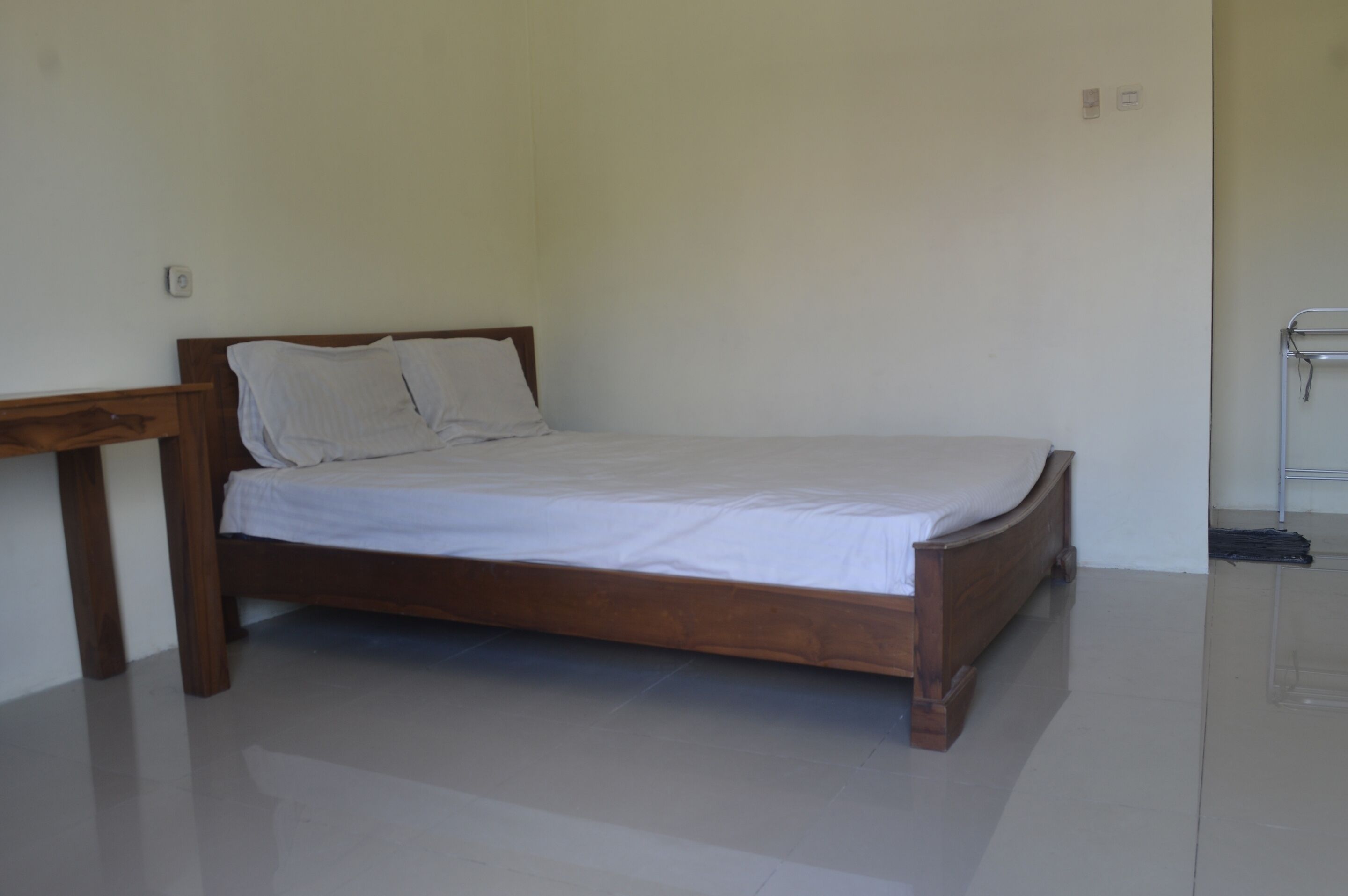 A single bed with a wooden frame and white bedding in a room with plain walls and a tiled floor.