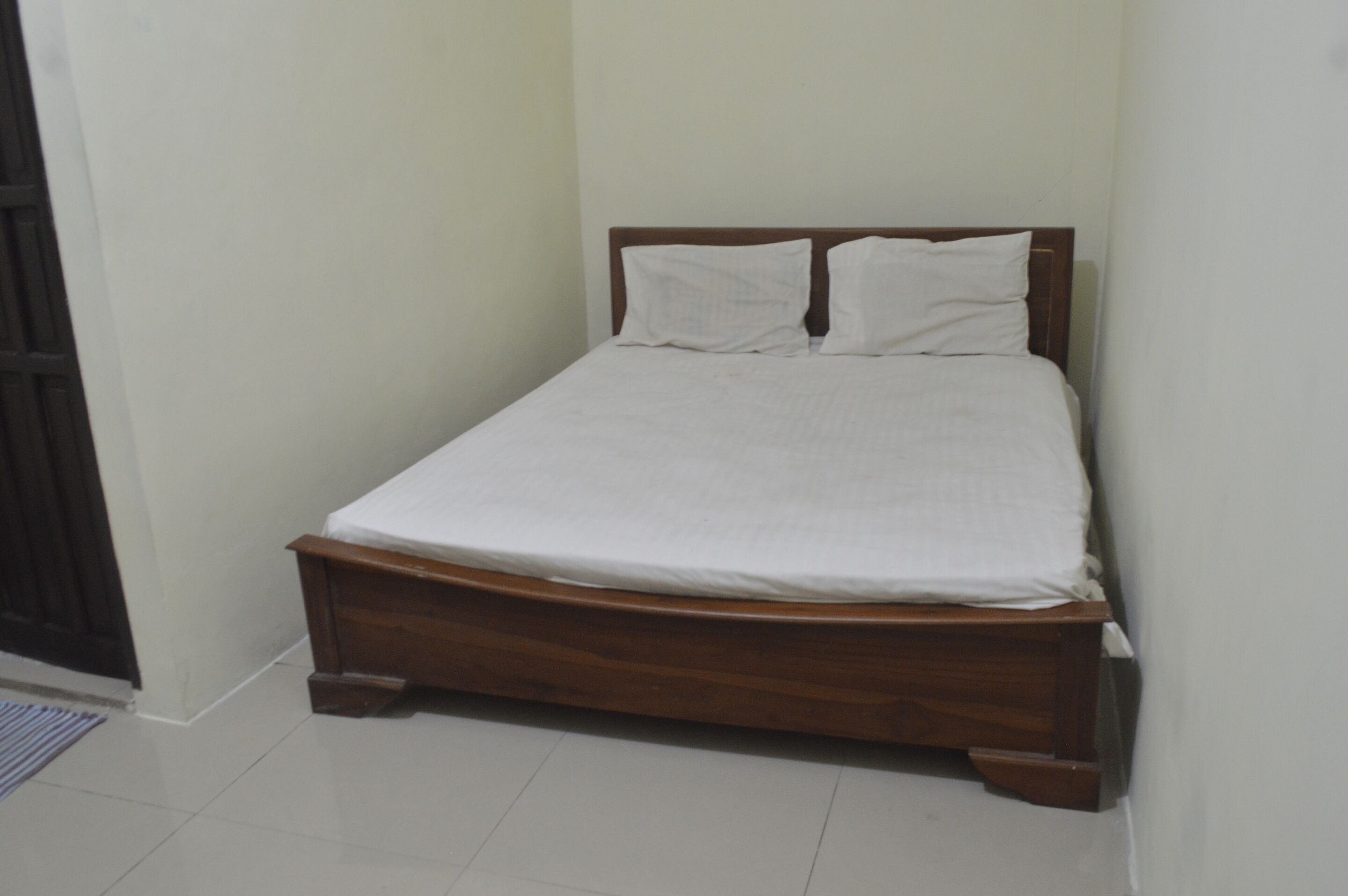 A single bed with a wooden frame and white bedding in a room with plain walls and a tiled floor.