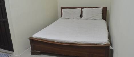 A single bed with a wooden frame and white bedding in a room with plain walls and a tiled floor.