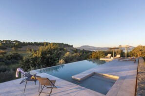 Un espace piscine extérieur moderne, doté de chaises longues et d’une pergola, entouré d’un paysage naturel et de collines.