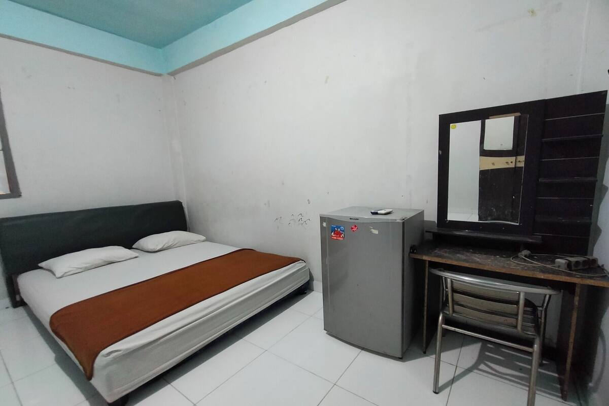 A small, simple room with a bed, a desk, a refrigerator, and a mirror.
