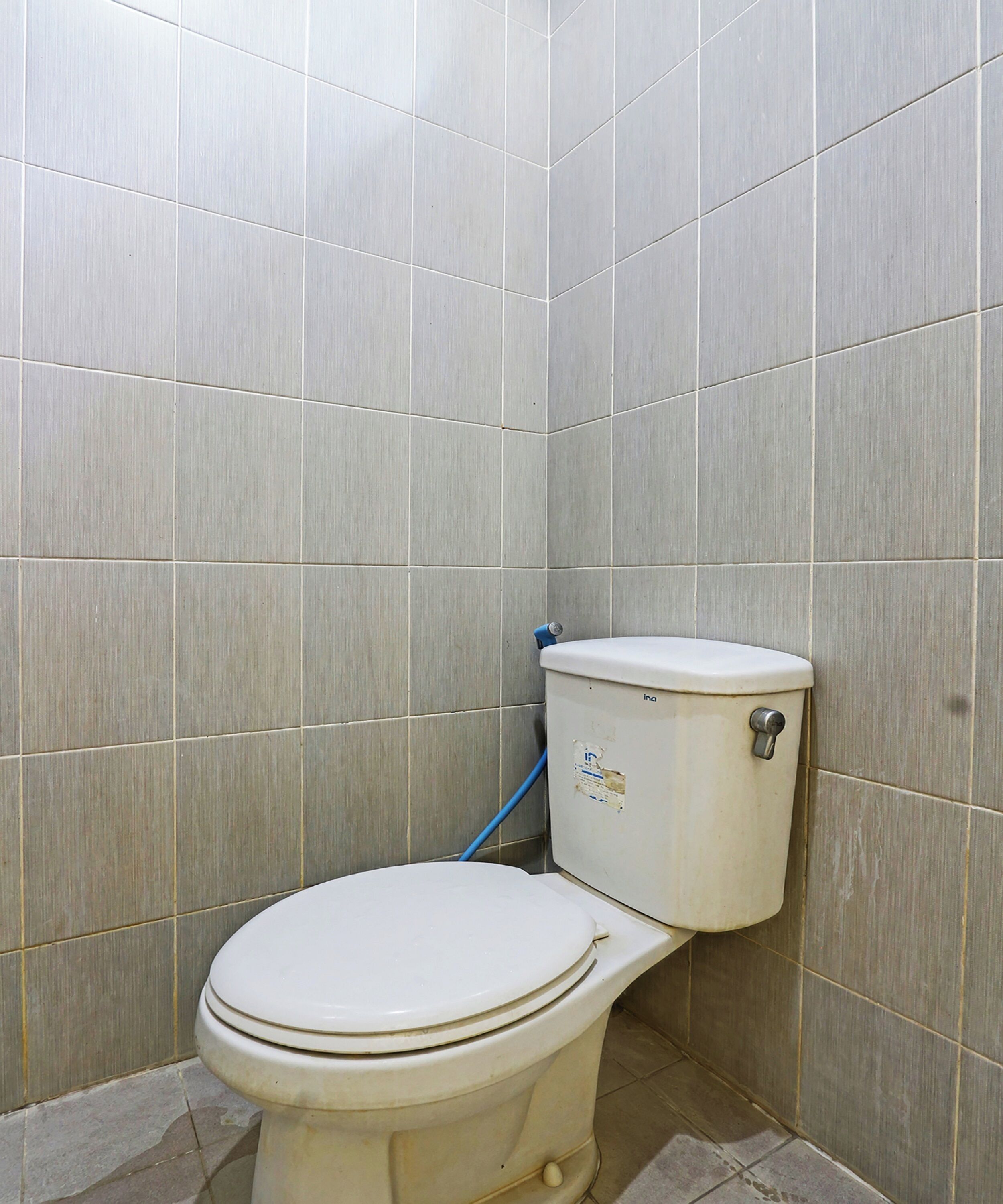 Ein kleines, gefliestes Badezimmer mit einer weißen Toilette und einem blauen Reinigungswerkzeug, das an der Wand lehnt.