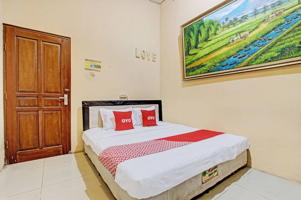 Une chambre avec une porte en bois, un lit avec une literie blanche et rouge, un tableau représentant un paysage et un panneau sur lequel est écrit le mot « AMOUR ».