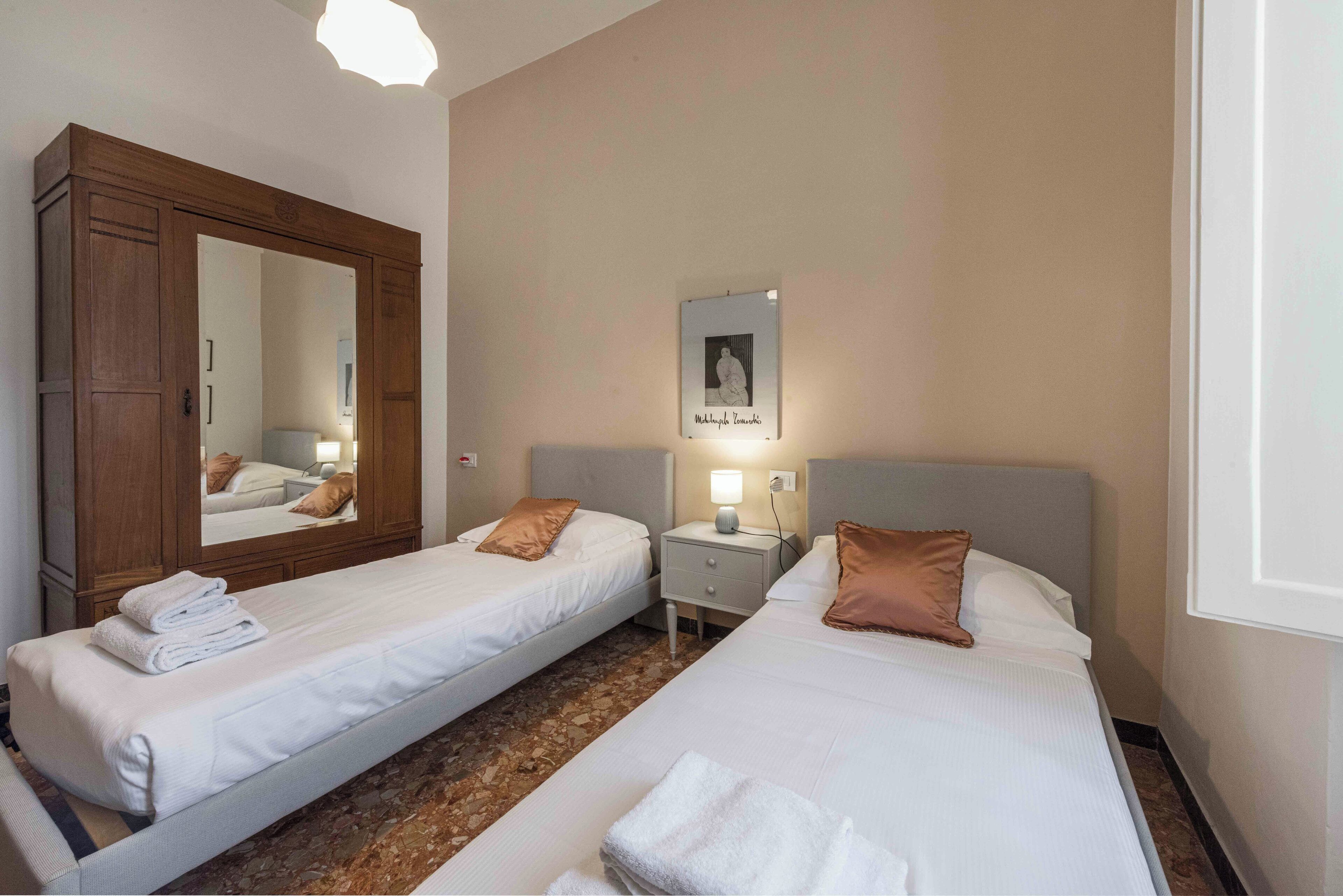 A hotel room with two single beds, a wooden wardrobe, a bedside table with a lamp, and a framed picture on the wall.