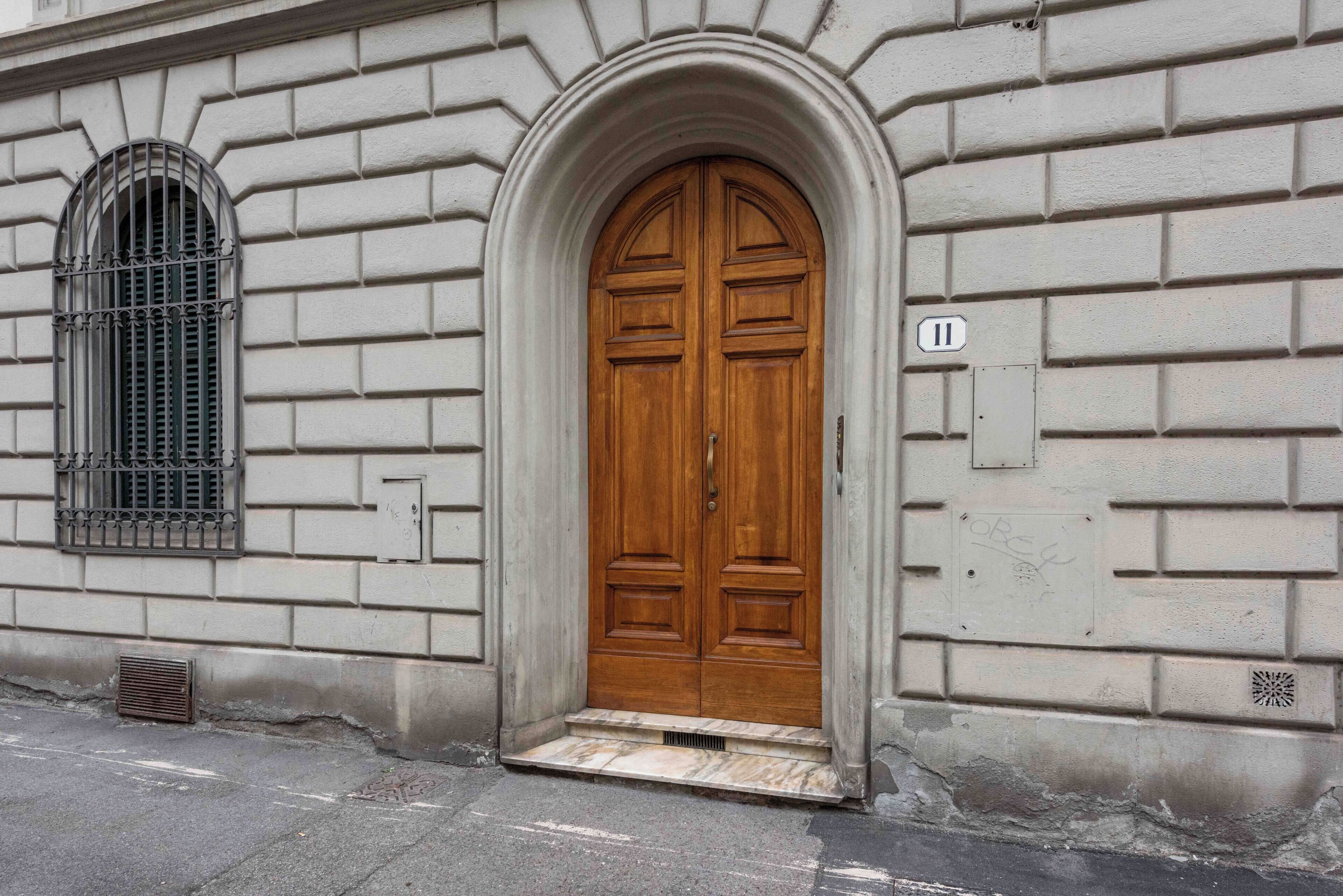 A large wooden double door with a small set of steps leading up to it, set in a building with a stone facade and a window with metal bars.