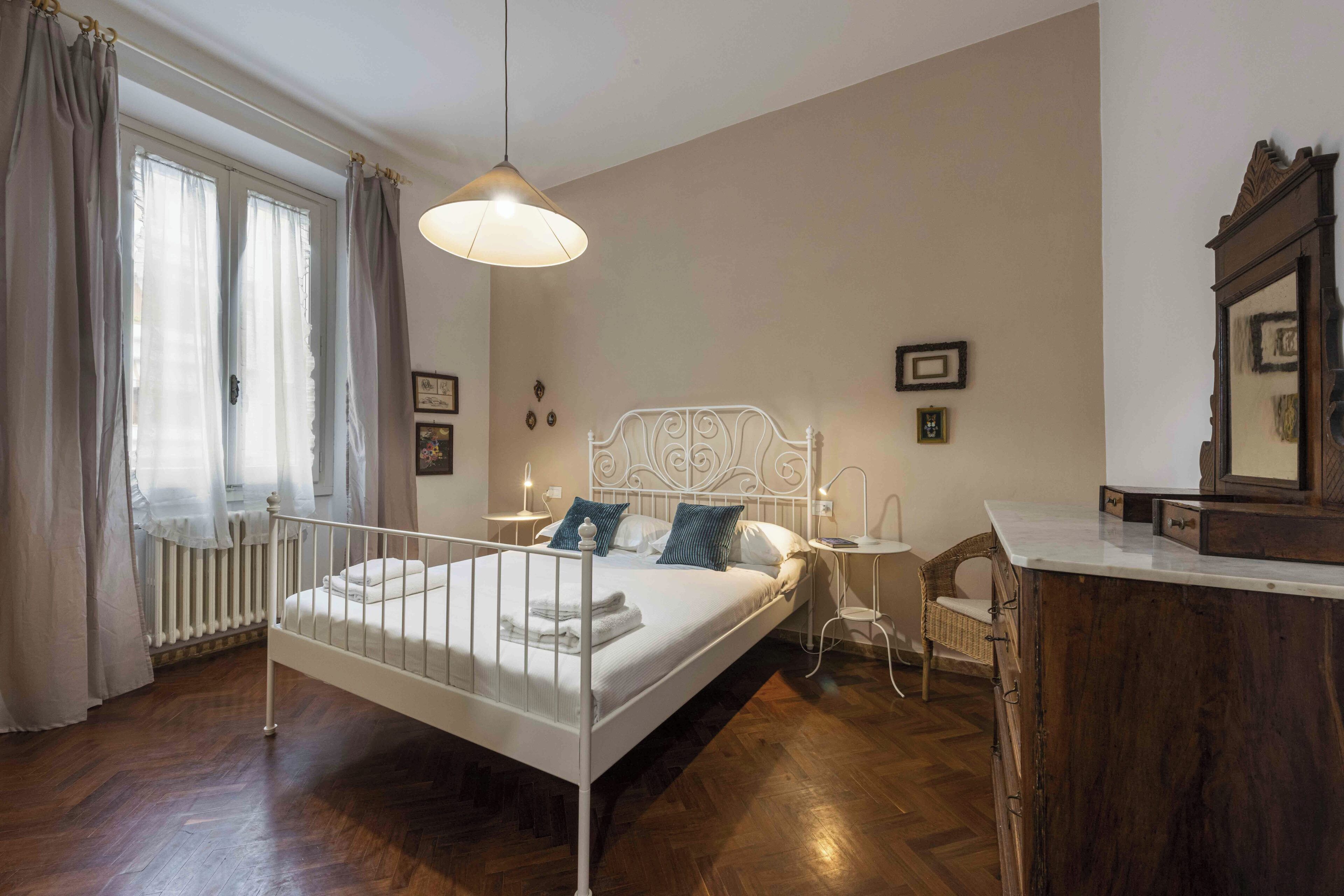 A bedroom with a white metal bed, a wooden dresser, a chair, and a window with curtains.