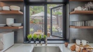 Een moderne keuken met een groot raam, inbouwplanken met servies en glazen, een spoelbak en een ontbijtbalk met gebak.