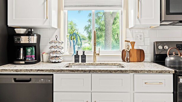 A kitchen with white cabinets, a granite countertop, a coffee maker, a kettle, and a window with a view of trees. - New! Comfortable 1BR Upper Flat, Roseville Downtown (Roseville)