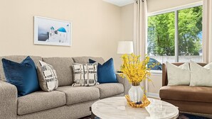 A living room with a beige sofa, a round coffee table, and a brown sofa. There is a window with a view of trees and a car outside. - Spacious 1BR End Unit, Heart of Roseville (Roseville)