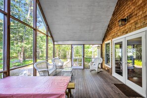 A covered porch with wicker furniture, large windows, and a view of the surrounding forest. - Water View Home With Great Screen Porch (2317) (Cotuit)