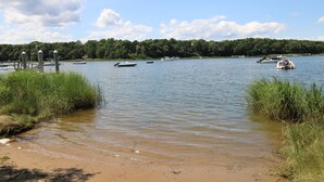 Un lago tranquilo con barcos, una orilla arenosa y una colina boscosa al fondo.