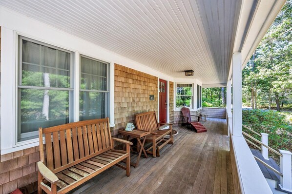 A covered porch with wooden furniture, large windows, and a view of the surrounding greenery. - Water View Home With Great Screen Porch (2317) (Cotuit)