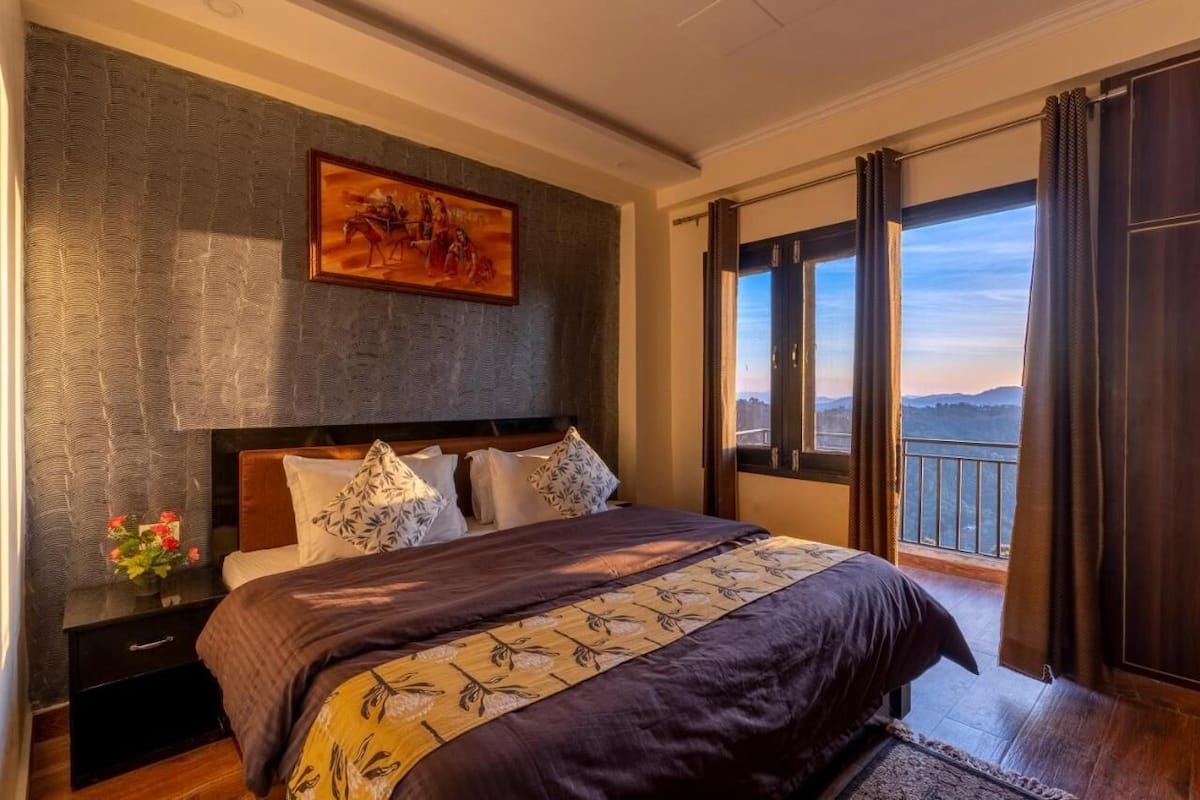 Une chambre à coucher avec un grand lit, une tête de lit, un tableau, une table de chevet, un vase avec des fleurs et une vue sur l’extérieur par une fenêtre.