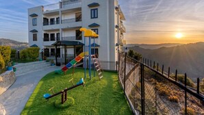 Un edificio de varios pisos con balcones, un parque infantil con tobogán y columpios, y vistas al atardecer sobre un paisaje montañoso.