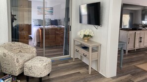 A modern living room with a glass door leading to a bedroom, a flat-screen TV mounted on the wall, a small wooden side table with a vase of flowers, and a patterned sofa. - Recently Updated & Complete w/Golf Cart, Coffee Bar, & MORE! (The Villages)