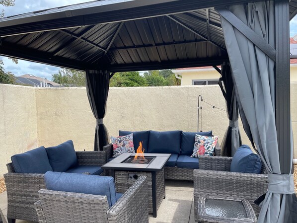 An outdoor patio area with a covered seating arrangement, a fire feature, and a pergola structure.