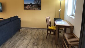 Una habitación con piso de madera, un sofá azul, una silla de madera con un cojín amarillo, una mesa de madera con una lámpara y un cuadro en la pared.
