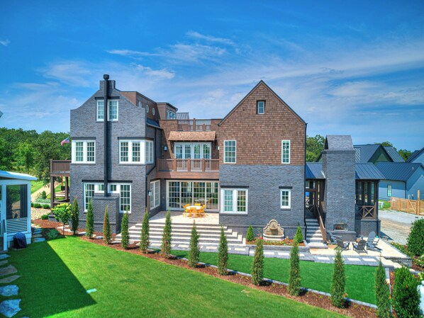 A large, multi-story house with a prominent stone facade, a spacious patio with outdoor furniture, and a well-maintained lawn.