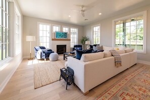 A spacious living room with a large sofa, a coffee table, and a fireplace. There are multiple windows providing natural light and a view of the outdoors.