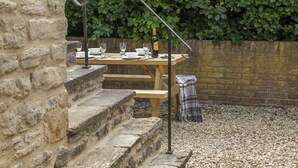 A wooden picnic table set up on stone steps with a brick wall and green foliage in the background. - The Milking Parlour - Luxury Cottages (Malmesbury)