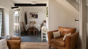A cozy living room with a brown leather sofa, a dining area with a wooden table and chairs, and a staircase leading to an upper floor. - The Milking Parlour - Luxury Cottages (Malmesbury)