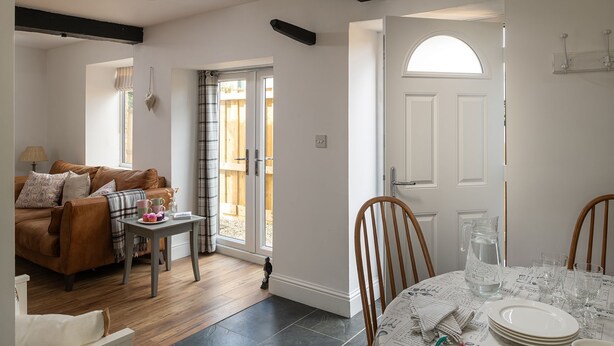 A cozy living room with a brown leather sofa, a small table with a lamp, and a view of a wooden deck through a glass door. - The Milking Parlour - Luxury Cottages (Malmesbury)