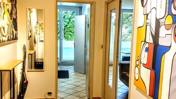 A hallway with a tiled floor, a vacuum cleaner, and a door leading to a room with a bed.