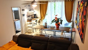 A living room with a sofa, a dining area with a table and chairs, a kitchen in the background, and a large window with curtains.
