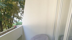 A balcony with a green artificial turf floor, a yellow umbrella stand, a folding chair, and a window with a view of trees.