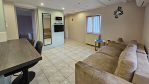 Un salon comprenant un canapé, une table à manger, un téléviseur fixé au mur et une fenêtre avec des rideaux.