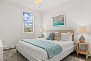 A bedroom with a bed, a window with blinds, a wall-mounted painting, two bedside tables with lamps, and a patterned rug. - Ash in the Sand | 30A | Privte Pool | Golf Cart | Bikes | Games (Santa Rosa Beach)