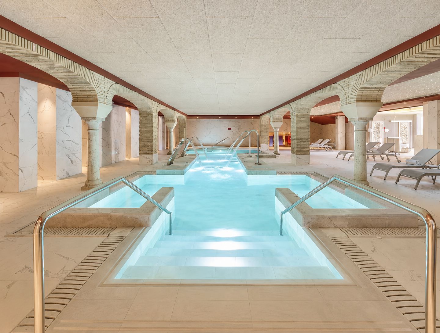 An indoor swimming pool with clear water, surrounded by marble walls and columns, and equipped with multiple entry points and lounge chairs.