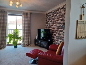 A living room with a red leather sofa, a television on a black stand, a potted plant, and a wall with a vintage-themed wallpaper. - Red Door Malt - Huku Kwetu (Dunstable) (Dunstable)