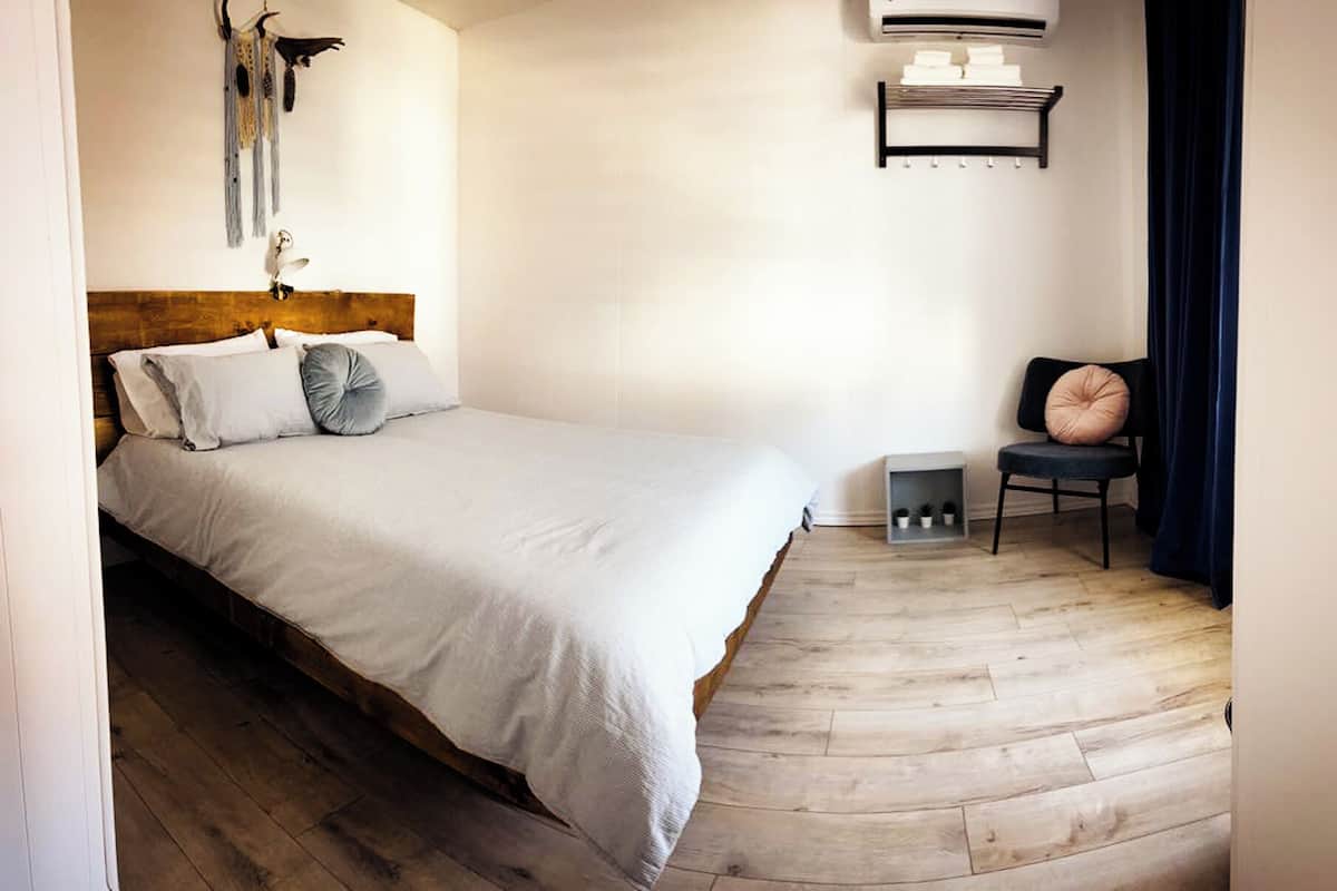 A bedroom with a wooden floor, a bed with white bedding, a chair with a cushion, a wall-mounted shelf, and a ceiling fan.