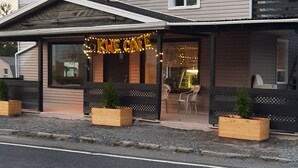 A two-story building with a cafe named "Kne Cafe" on the ground floor. There are outdoor seating areas with wooden planters and a satellite dish on the roof. - KWE Café & Auberge (Pohenegamook)