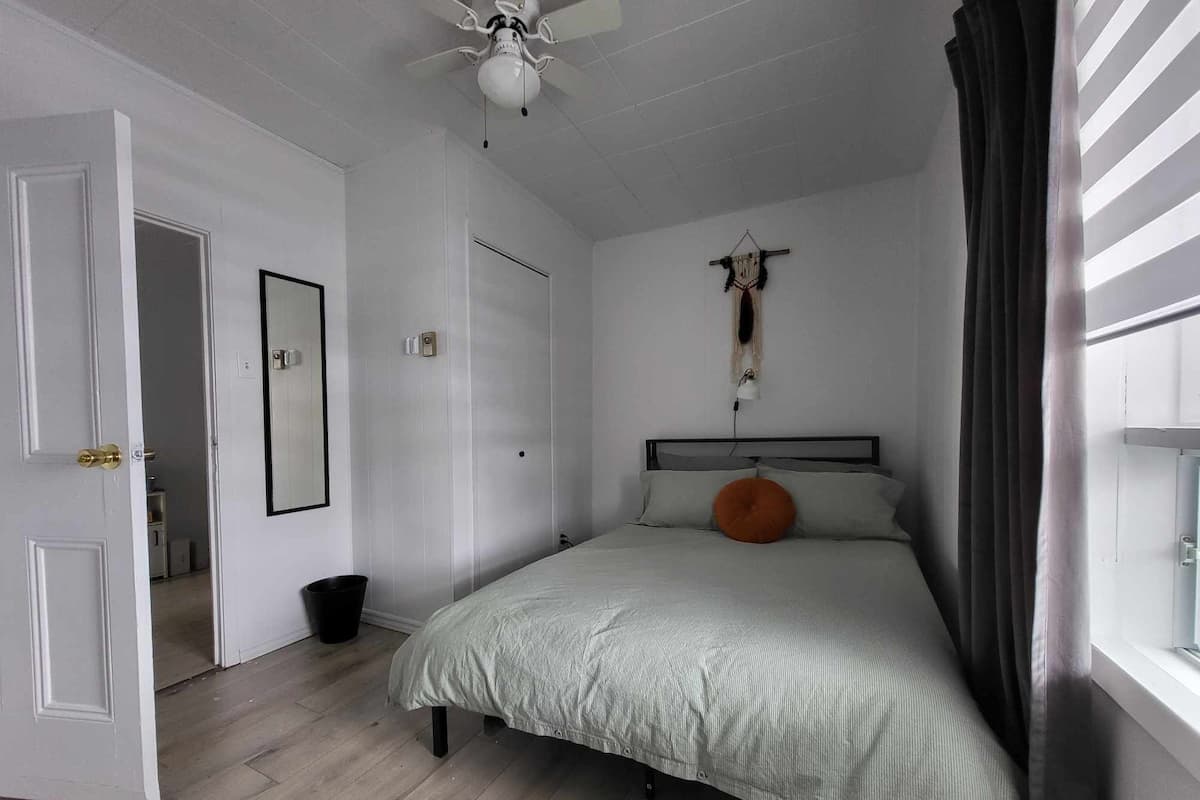A bedroom with a bed, a ceiling fan, a wall-mounted light, a door, and a window with curtains.