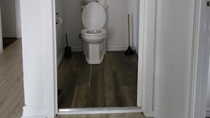 A small, white-walled bathroom with a toilet, a broom, and a toilet brush. - KWE Café & Auberge (Pohenegamook)