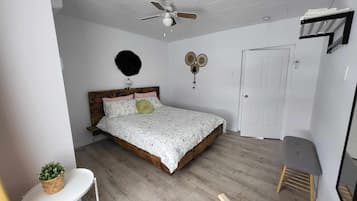 Une chambre avec un lit en bois, un ventilateur de plafond, une petite table avec une plante et un banc.