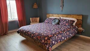 A bedroom with a wooden ceiling, a large bed with floral bedding, a side table, a hanging light, and a window with curtains. - KWE Café & Auberge (Pohenegamook)