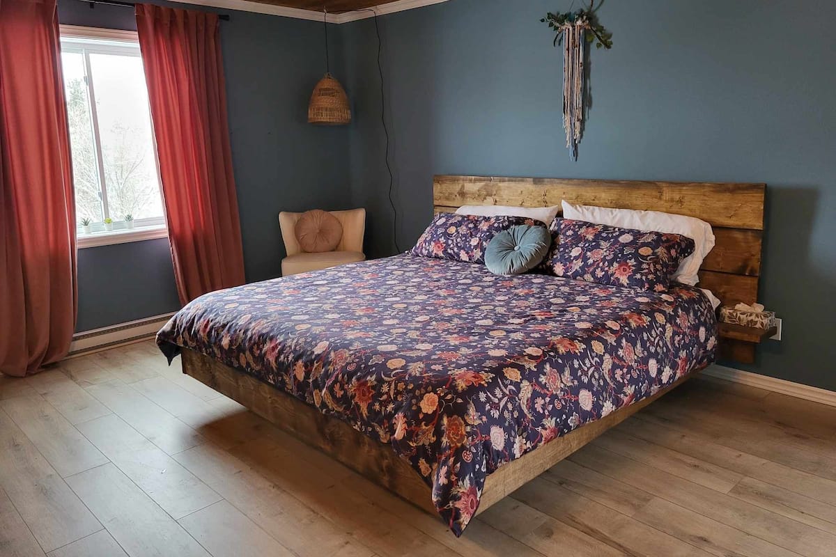 A bedroom with a wooden ceiling, a large bed with floral bedding, a side table, a hanging light, and a window with curtains.