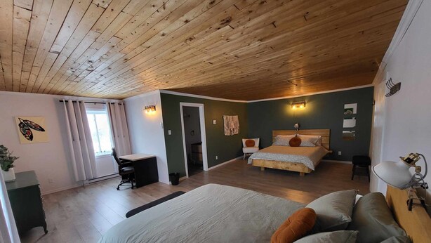 A bedroom with a wooden ceiling, a large bed, a desk, and a chair. - KWE Café & Auberge (Pohenegamook)