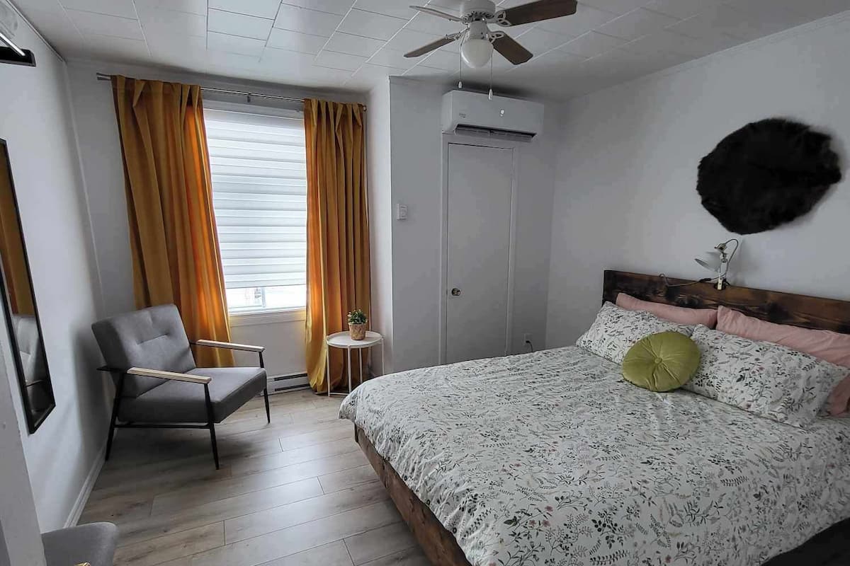 A bedroom with a bed, a chair, a ceiling fan, a window with curtains, and an air conditioning unit.