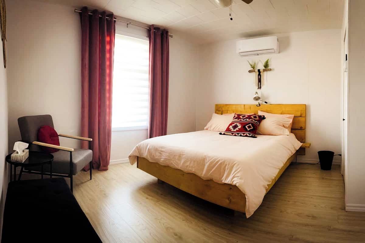 A bedroom with a wooden bed, a chair, a bedside table, a window with curtains, an air conditioning unit, and a wall-mounted plant.