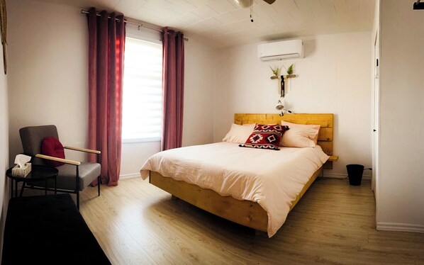 Une chambre avec un lit en bois, une chaise, une table de chevet, une fenêtre avec des rideaux, un climatiseur et une plante fixée au mur.