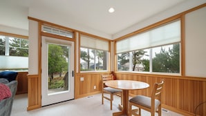 A room with a wooden paneled wall, a sliding glass door leading to a balcony, a round wooden table with chairs, and a grey sofa with a red blanket.